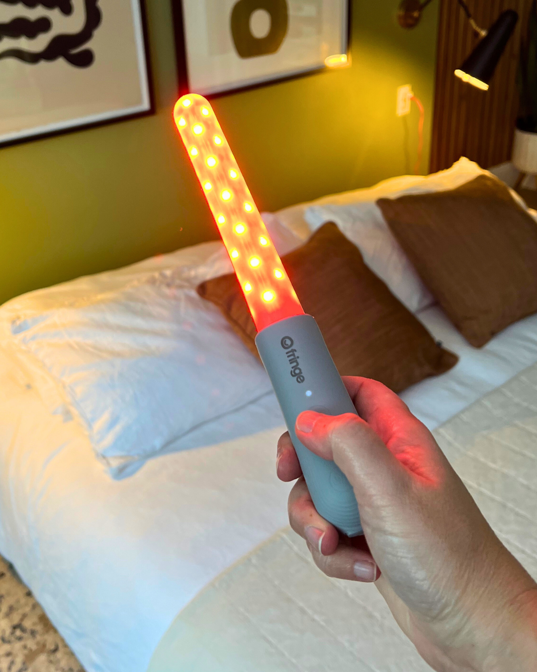 Hand holding the Fringe Heals pelvic floor wand lit red on a bed
