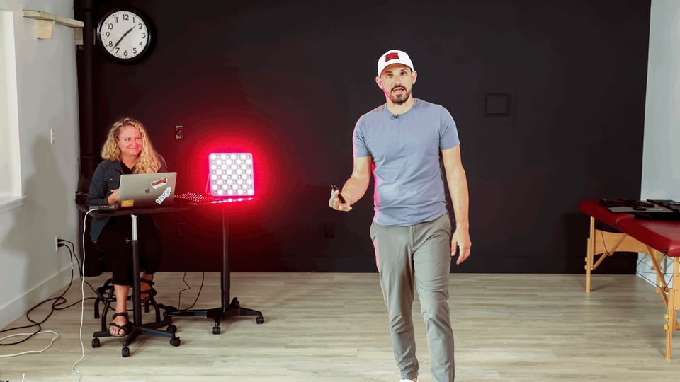 A man demonstrates red light therapy with a Fringe LED panel while a woman operates a laptop.